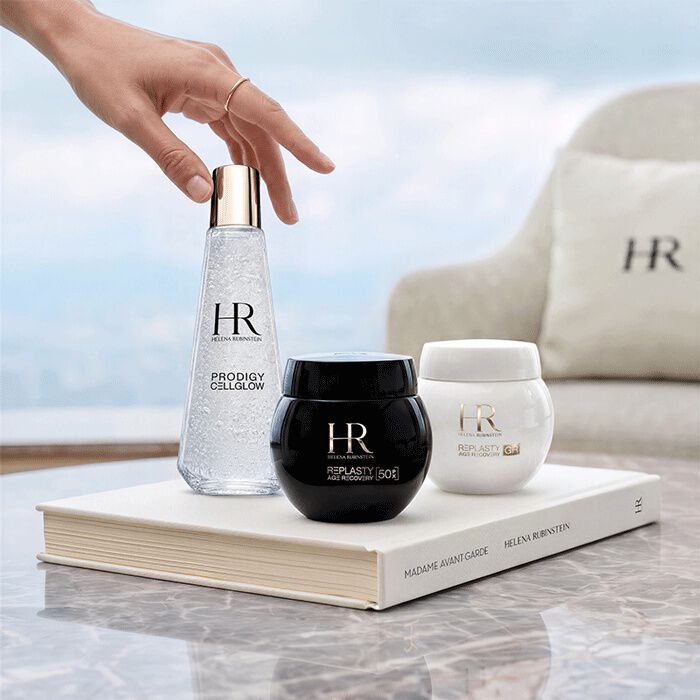 Three Helena Rubinstein skincare products (Prodigy Cellglow and Re-Plasty) displayed on a white book in front of a window with a landscape view.​