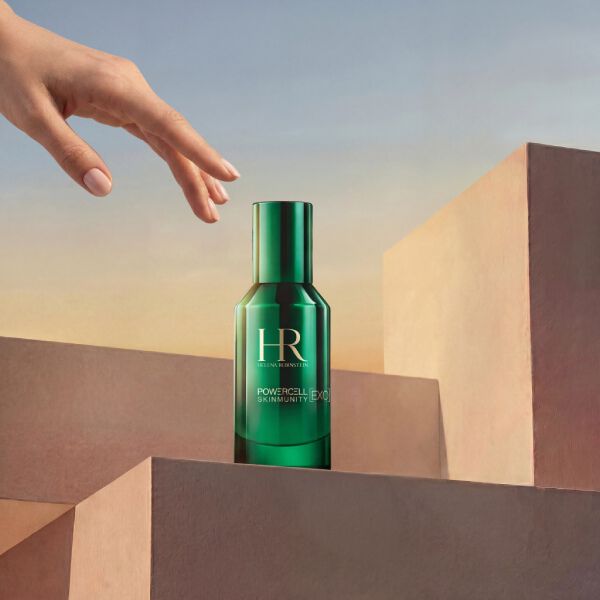 Product shot of Helena Rubinstein Powercell Skinmunity Exo Serum in a glossy green glass bottle. The bottle sits atop a stack of pink geometric podiums, with an elegant hand reaching down towards it from above, against a warm sunset-toned sky background.​