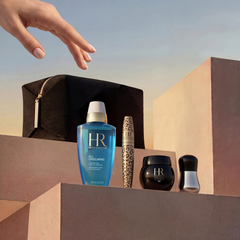 An elegant hand reaches down toward a curated selection of Helena Rubinstein products arranged on minimalist, sandy-beige stone blocks. The set is displayed against a warm, golden-hour sunset sky and features a black travel pouch, the blue 'All Mascaras' eye makeup remover, a leopard-print Lash Queen Feline Blacks mascara, a small black Re-Plasty cream jar, and a silver applicator tool.​