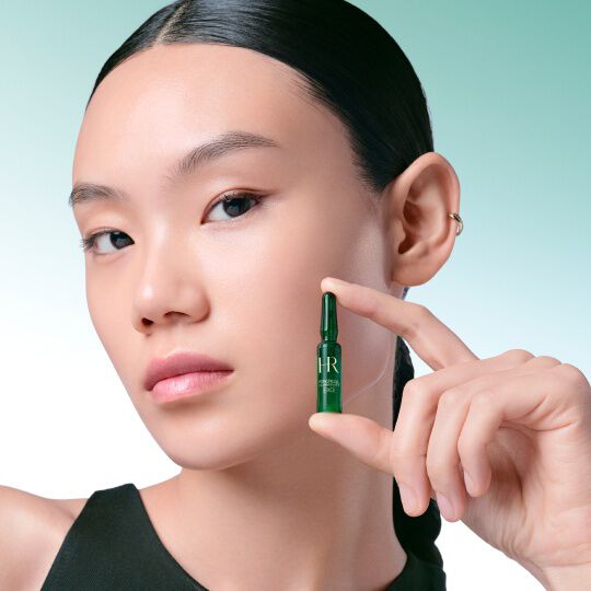 Close-up of a woman's face delicately holding a green Helena Rubinstein Powercell [EXO] Skinmunity ampoule between her fingers, against a green and white gradient background.​