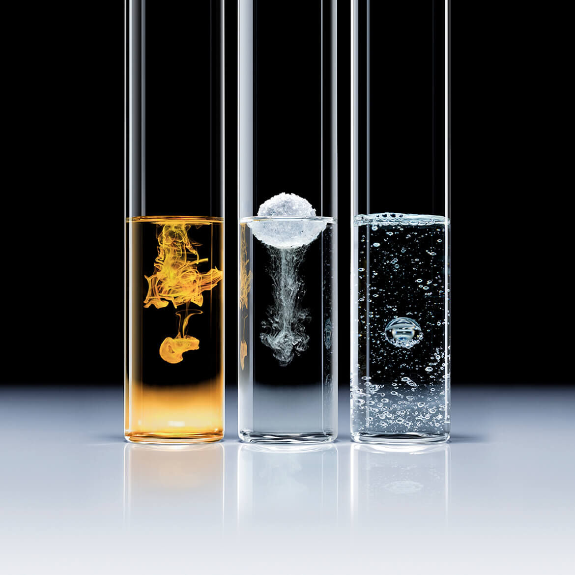 Three glass test tubes containing different liquids: the first a yellow liquid with a suspended drop, the second a clear liquid with a dissolving solid, and the third a sparkling liquid with a bubble.