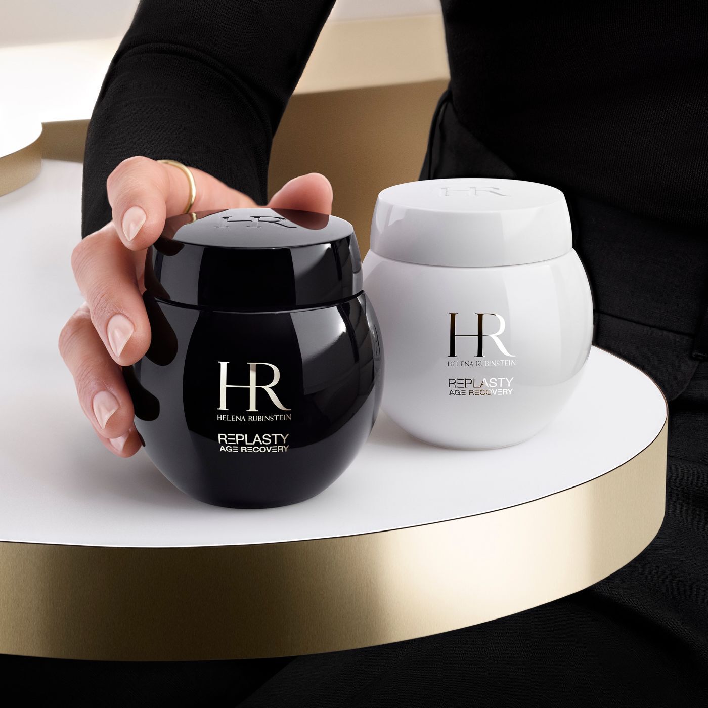 A hand grasps a black Helena Rubinstein Re-Plasty Age Recovery cream jar, with a matching white jar beside it, on a golden platform.