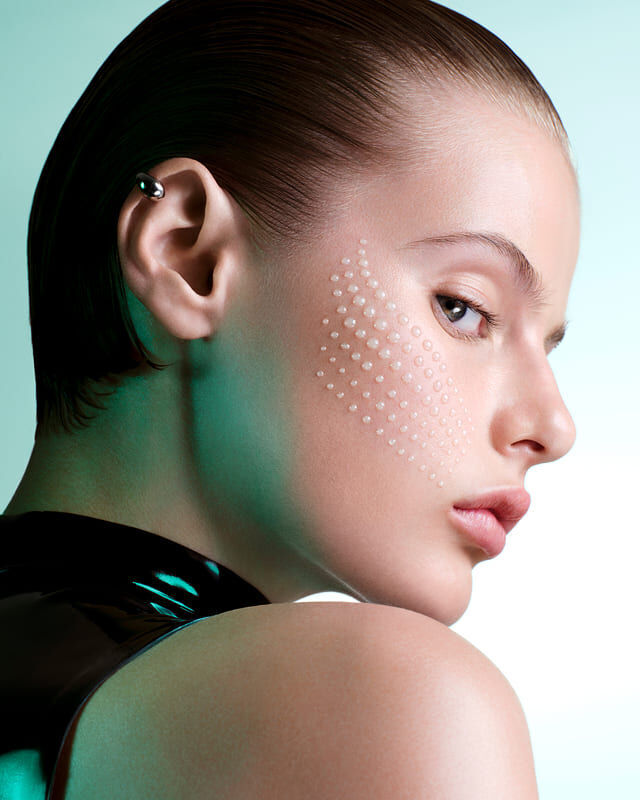 a woman posing in profile with drops of serum on her cheek against a blue background