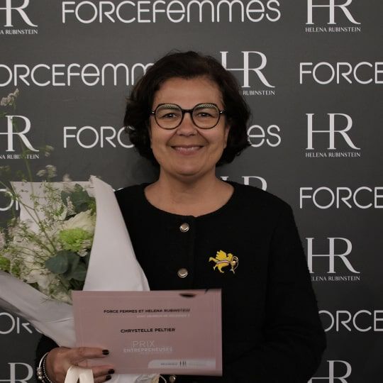 A smiling woman with short, dark hair, wearing glasses and a purple jacket, holds a bouquet of white flowers in front of a logo-covered wall.