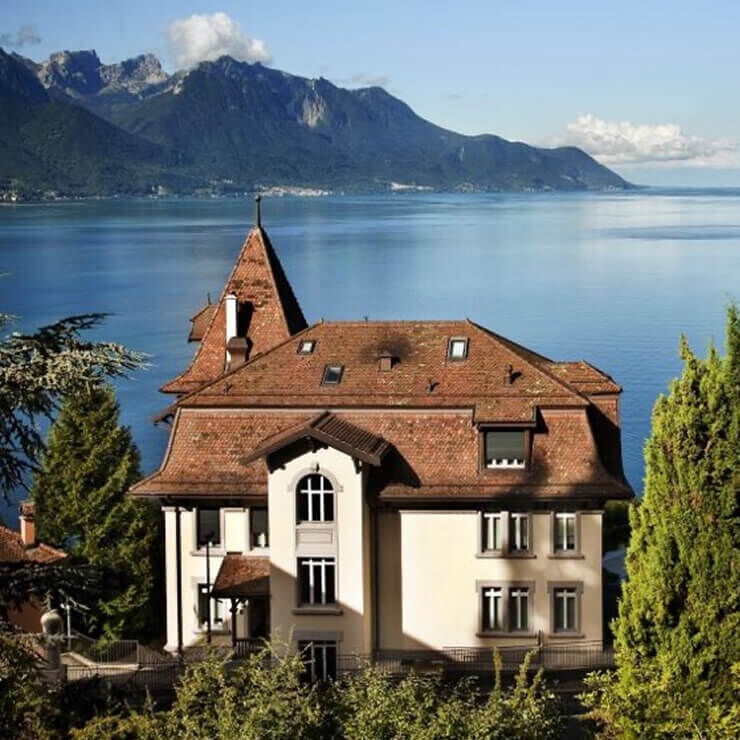 A large traditional-style house with a red-tiled roof and light-colored walls, situated by a large deep blue lake, surrounded by mountains. The sky is clear.