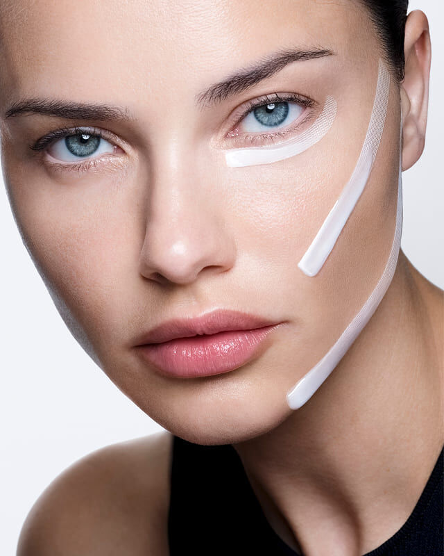 Adriana Lima with white cream and strips applied to her face, demonstrating a visible lifting effect.