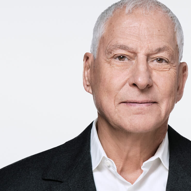 Close-up of an elderly man with gray hair, white shirt, and dark jacket, looking slightly to the side. His expression is neutral and his face is illuminated.