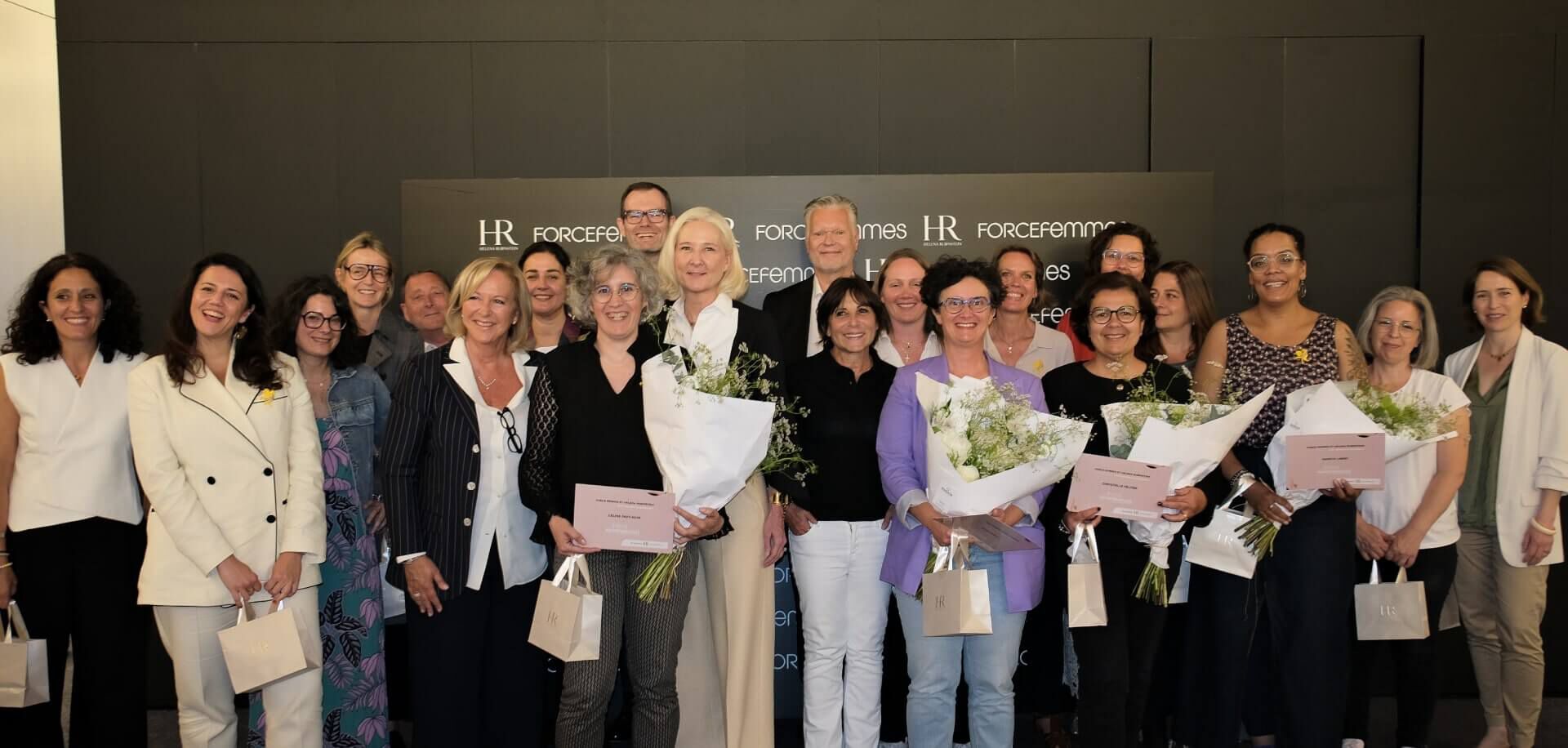 Large group of people, mostly women, some holding bouquets and bags, posing cheerfully in front of a black wall with "HR" and "FORCEfemmes" logos.