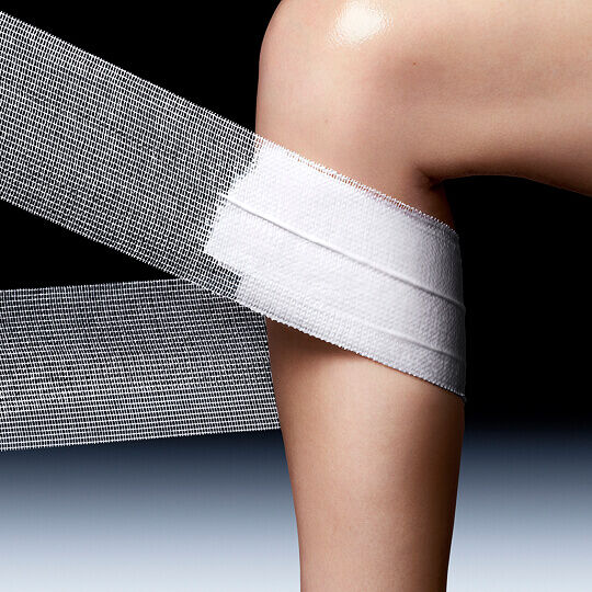 A knee partially covered with a wide, textured white mesh band, against a dark background, evoking care or a bandage.