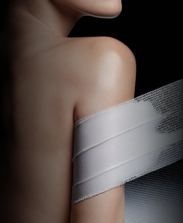 Bare shoulder and arm partially covered with a white mesh band, suggesting skin care or treatment, against a dark background.