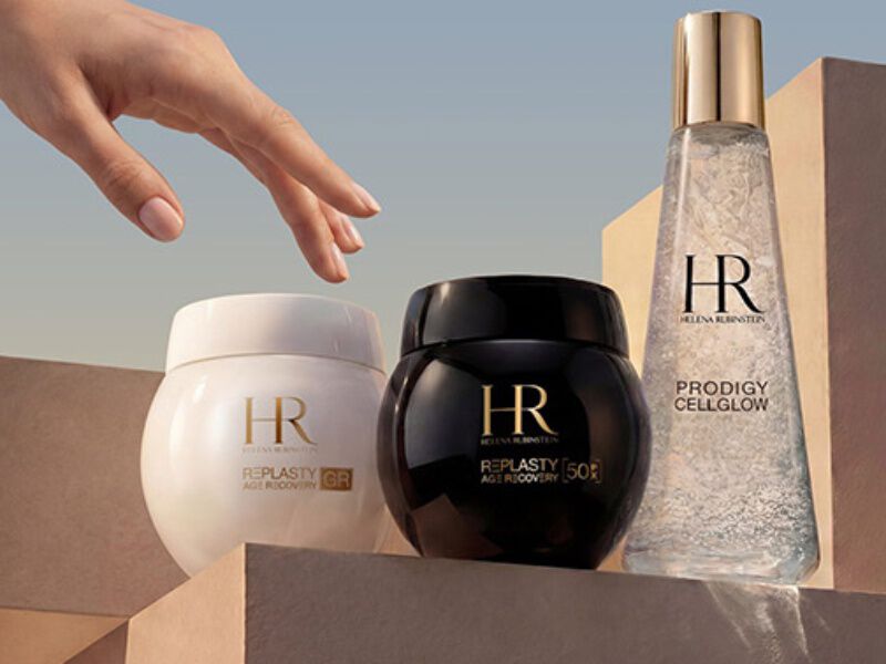 A hand extends towards three Helena Rubinstein products, showcasing an elegant design and soft lighting.