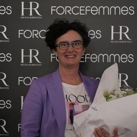 A smiling woman with short, dark hair, wearing glasses and a purple jacket, holds a bouquet of white flowers in front of a logo-covered wall.