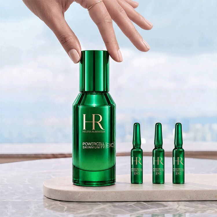A hand touching the green bottle of Helena Rubinstein Powercell [EXO] Skinmunity serum, placed next to three [EXO] Shots ampoules on a marble stand against a bright landscape view.​