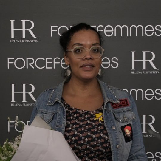 A smiling woman with dark, tied-back hair, wearing round glasses and a denim jacket, holds a bouquet of white flowers in front of a logo-covered wall.