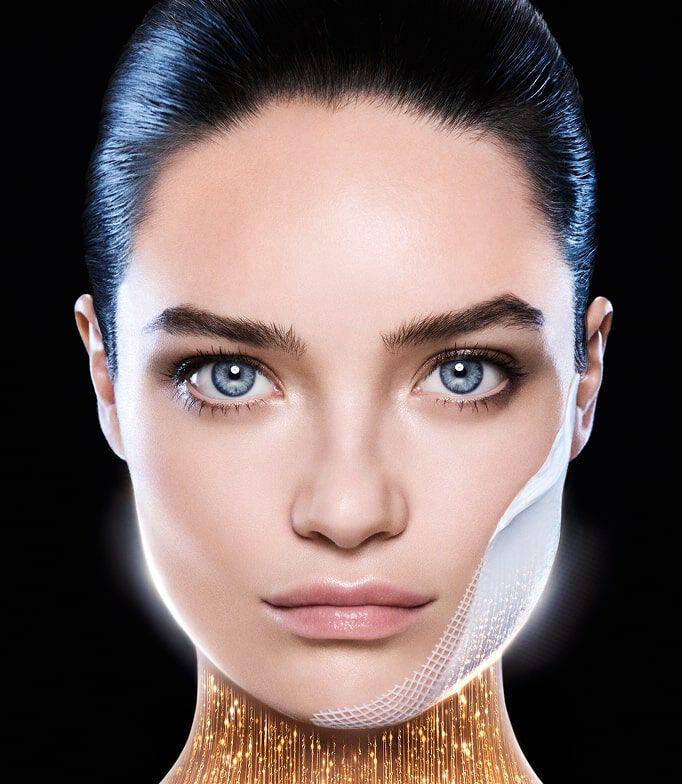 Woman face with blue eyes, a fine patch on her cheek. Below her chin, a golden glow symbolizes a lifting or revitalizing effect. Black background.