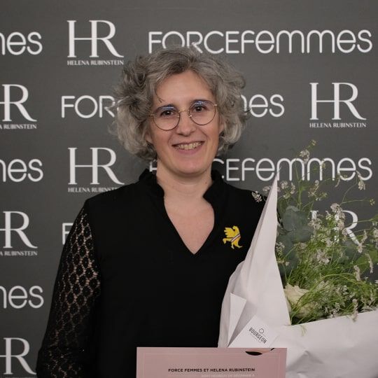A smiling woman with curly gray hair and glasses holds a bouquet of flowers. She wears a black top with lace detail. Logo background.