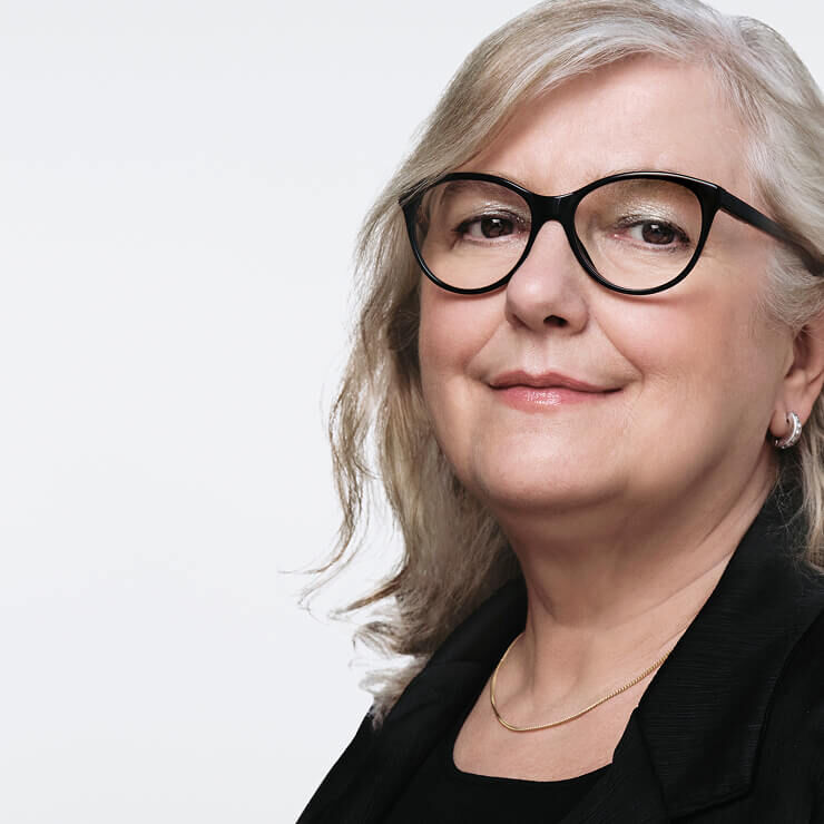 Close-up of a middle-aged woman with light gray hair, wearing black glasses and a subtle gold necklace, smiling slightly.